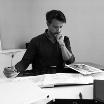 Man seated at a light surface with scattered papers and graphics, holding a pen and wearing a dark long-sleeved shirt