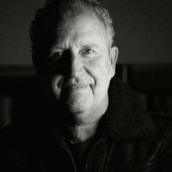 Grayscale portrait of a man with short textured hair wearing a dark jacket, looking forward with a slight smile against a blurred dark background