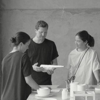 Three people gather around a light surface displaying small white bowls and cylindrical shapes in grayscale