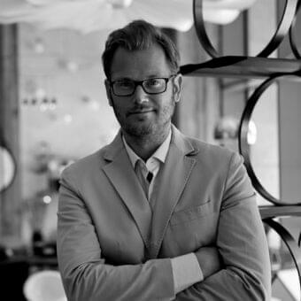 Man in light suit with crossed arms and glasses centered against blurred circular metal and architectural shapes in grayscale