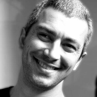 Close-up black and white portrait of a smiling man with short textured hair and stubble, angled right with gradient gray background