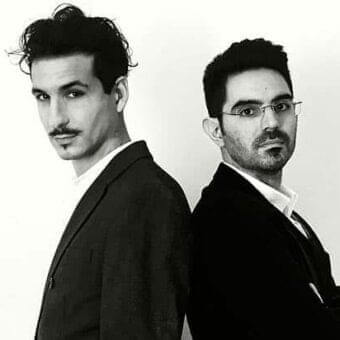 Two men in jackets and collared shirts stand back to back against a light gray monochromatic background