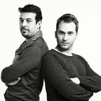 Two men with crossed arms stand facing forward against a light gray monochrome background