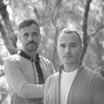 Two men in light jackets stand close together before a blurred background of trees and dark foliage