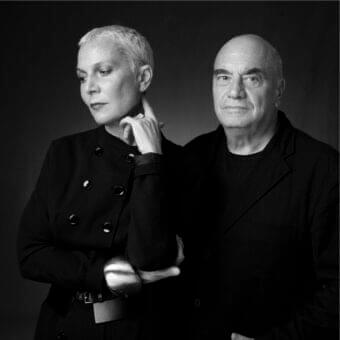 Monochrome portrait shows two people facing forward with dark outerwear and short hair against a dark background
