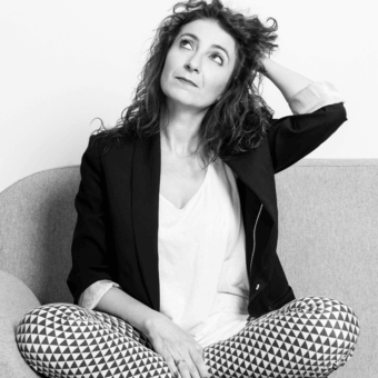 Woman with curly hair seated on a curved light surface looking upwards with a raised hand wearing patterned trousers