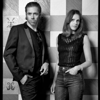 Monochromatic image of two people standing against a background of large gray squares with indistinct dark graphic details