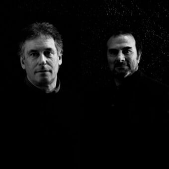 Black and white portrait of two men in dark jackets against a dark concrete texture, featuring subtle “studio kairos” text.