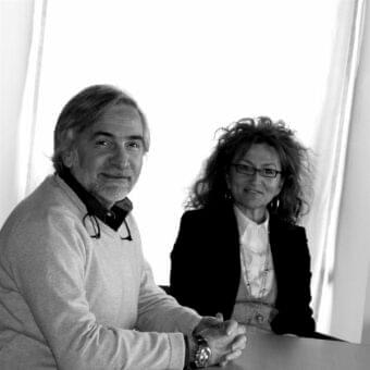 Grayscale portrait of two people facing forward, one man with gray hair and one woman with dark curly hair, illuminated from the right