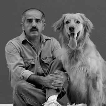 Seated man in denim clothing and a large dog with open mouth are shown in grayscale against a plain gray background