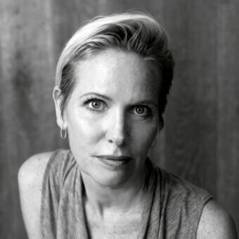 Black and white portrait of Terri Pecora with short hair, hoop earrings, and a sleeveless draped top against a textured backdrop.