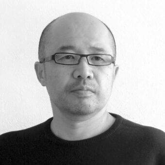 Portrait of Toshuyuki Yoshino, a bald man with glasses, wearing a black sweater, against an off-white wall with diffused lighting.