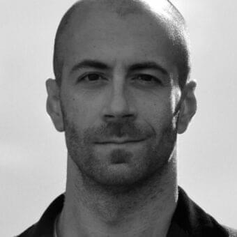 Monochrome portrait of a man with a shaved head and facial stubble looking forward against a light gray background