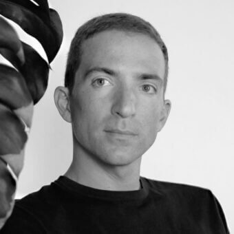 Black and white portrait of Valerio Sommella, a man with short dark hair, wearing a black t-shirt, framed by partial Monstera leaves.