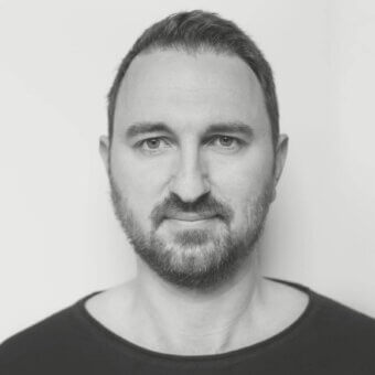 Grayscale portrait of a man with short dark hair and a full beard wearing a dark garment against a light gray background