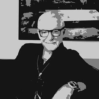 Seated person with glasses and dark clothing faces forward against a large rectangular patterned background
