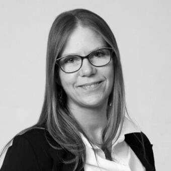 Gray scale portrait of a woman with medium brown hair wearing glasses and a dark jacket while smiling forward