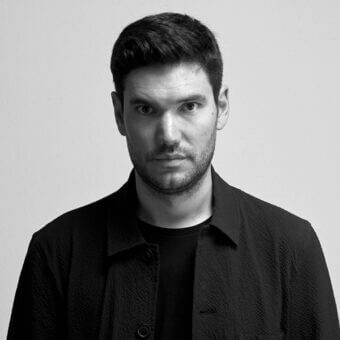 Man in black double-breasted jacket, dark hair, short beard, neutral gray background, portrait with direct gaze, textured fabric visible.