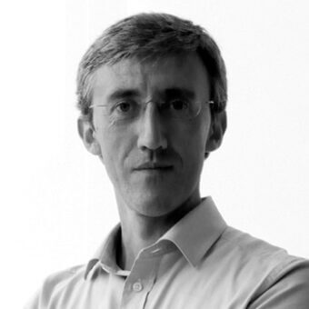 Black and white portrait of Alessio Bassan, facing forward with short gray hair, wearing glasses and an open-collar shirt, on a white background.