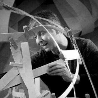 Person with dark hair stands within a grayscale web of curved light-colored strips and angular connections