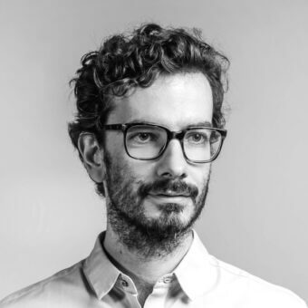 Black and white portrait of Andrea Steidl with glasses, short curly hair, and a light button-down shirt against a gray gradient backdrop.