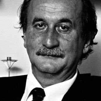 Man in dark suit and tie looks forward with mustache, framed by dark hair and shadowed facial features against a light backdrop