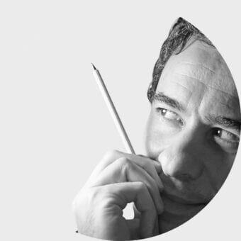 Black and white portrait of Angelo Tomaiuolo in circular frame, holding a pencil to his cheek against a light gray backdrop.