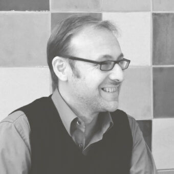 Black and white portrait of Antonio Rodriguez, wearing a vest and glasses, set against a geometric tiled wall backdrop.