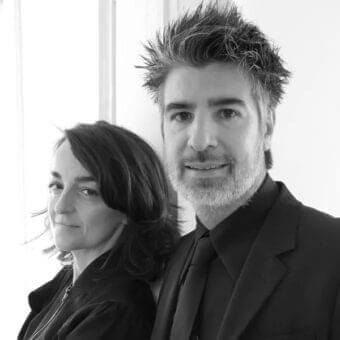 Two people are shown in close-up, a man with beard in a suit and a woman with dark hair, facing forward against a light backdrop