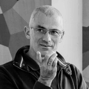 Man with short hair and glasses looks forward with hand to chin against a gray geometric patterned wall