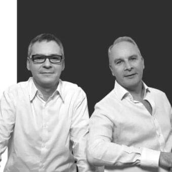 Two men in light shirts stand close together against a divided grayscale background with contrasting tones