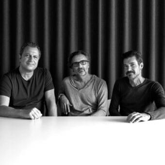 Three men stand behind a light horizontal surface with hands resting on top against a dark ribbed backdrop