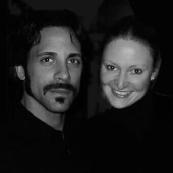 Black and white portrait of a man and woman, dark clothing, neutral and smiling expressions, indistinct textured background, high contrast.