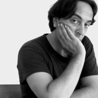 Black and white portrait of a man with dark hair in a contemplative pose, wearing a dark t-shirt against a light gray background.