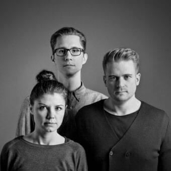Three people face forward against a dark gray backdrop with neutral expressions and similar coloring