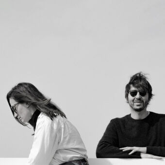 Black and white portrait of a woman and man flanking a white surface, featuring contrasting attire and direct gaze.