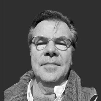 Middle-aged man with short hair and glasses looks downward wearing a cable knit garment against a dark gray background