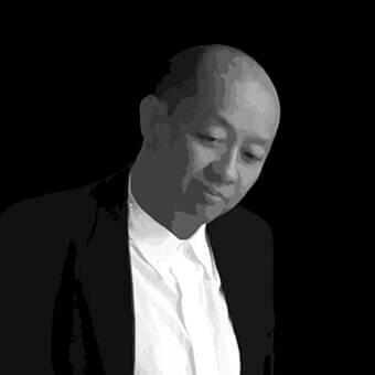 Grayscale portrait of a person with a shaved head wearing a collared shirt and dark jacket with a loosely knotted tie
