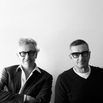Two people with glasses gaze forward wearing dark jackets and sweaters against a gray monochrome background