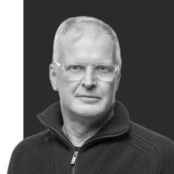 Grayscale portrait of a man with short hair and glasses wearing a dark cable knit top against a divided dark and white background