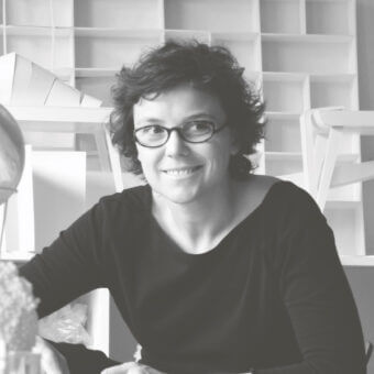 Constance Guisset portrait, black and white, short curly hair, glasses, architectural shelving backdrop, soft diffused lighting.