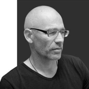 Monochromatic portrait of a man in profile with glasses, beard, and dark v-neck garment against a two-tone gray background