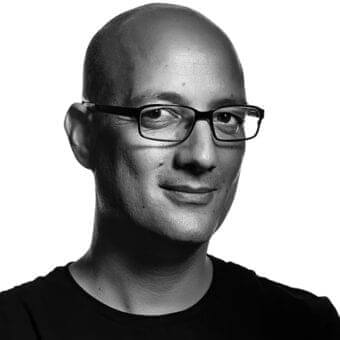Grayscale portrait shows a smiling person with a shaved head and rectangular glasses wearing a dark, rounded-neck garment against a white background