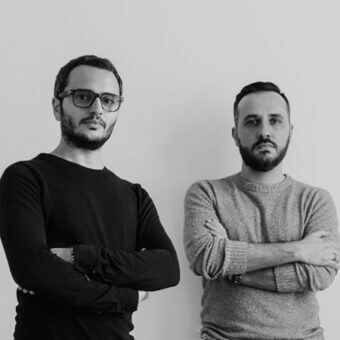 Black and white portrait of two men with crossed arms, wearing crew neck sweaters and glasses, against a plain white backdrop.