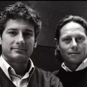 Black and white portrait of two men, edi and paolo ciani, with dark sweaters, neutral expressions, against a dark textured backdrop.
