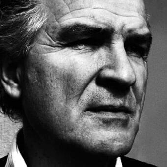 Close-up monochrome portrait of an older man facing downward with visible wrinkles and graying hair against a gray background
