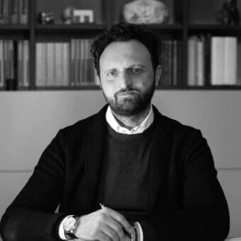 Man with dark curly hair and beard holds a dark writing tool before grayscale bookshelves filled with books and objects