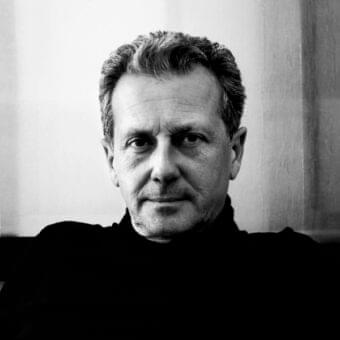 Grayscale portrait of a man facing forward with short gray hair and visible facial lines wearing a dark high-necked garment