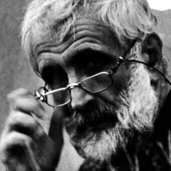 Close-up black and white portrait of a mature person with facial hair, hand raised partially obscuring the face and wearing eyeglasses