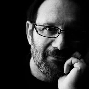 Man with beard and glasses in close-up, hand raised to mouth, shadowed face, dark background